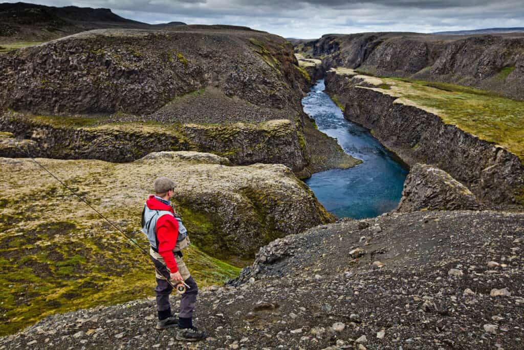 Fishing enthusiast overlooking a canyon with a river in Iceland, emphasizing freshwater fishing and scenic landscapes.