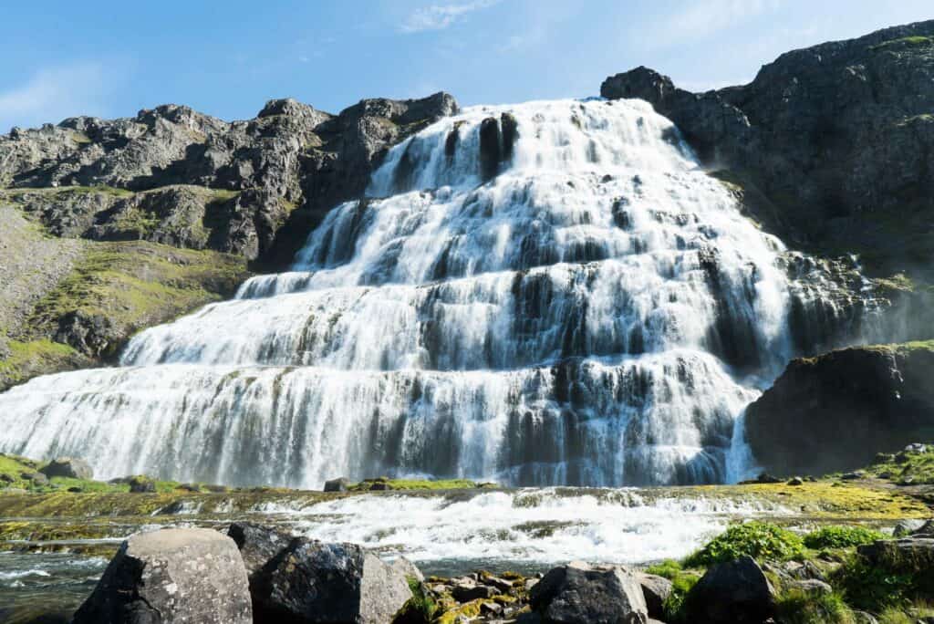 Vibrant waterfall cascading over rocky terrain with lush greenery and blue sky, ideal for fish farming and aquaculture tourism.