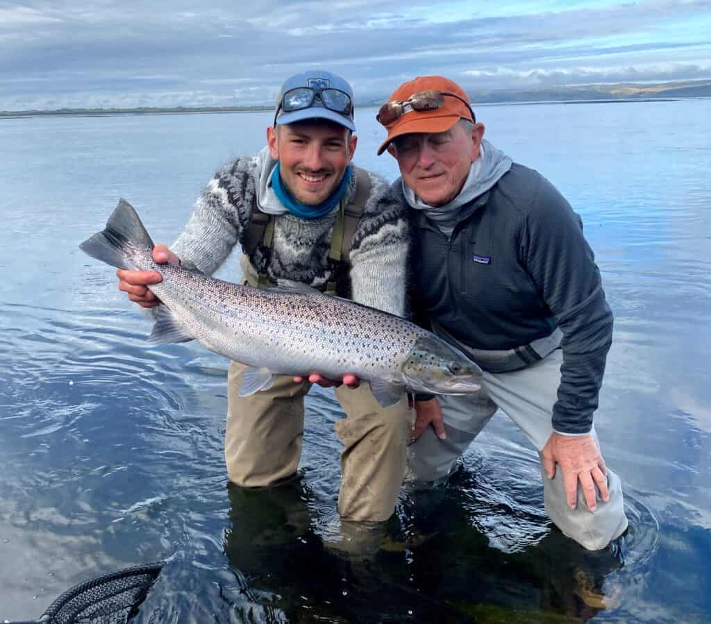 Two anglers holding a large fish in a river with cloudy sky.