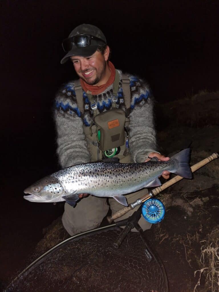 Man holding a large sea trout at night with fishing gear.