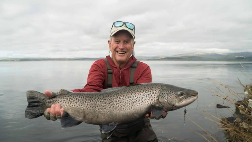Fisherman holding a large brown trout by a river.