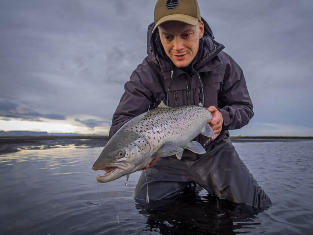 Man holding a large sea trout over water with cloudy sky.