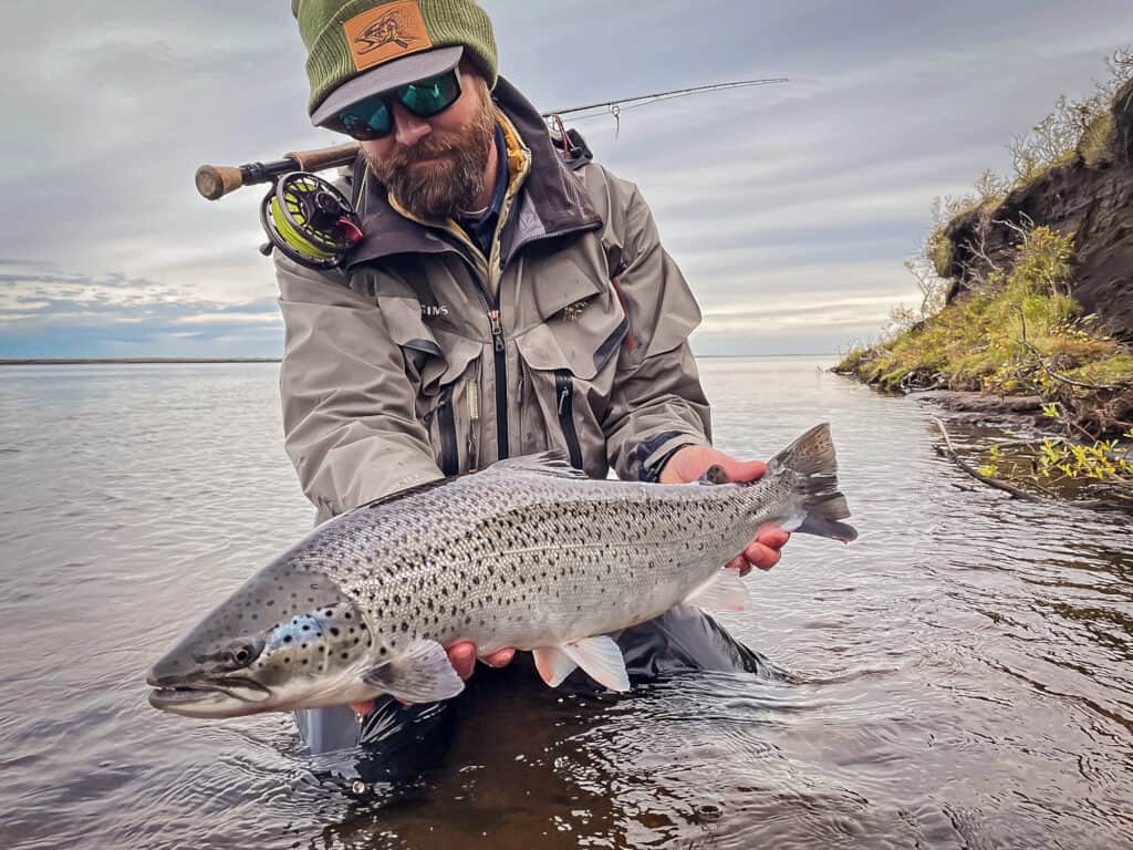 Fisherman holding a sea trout in a river with a fishing rod on his back.