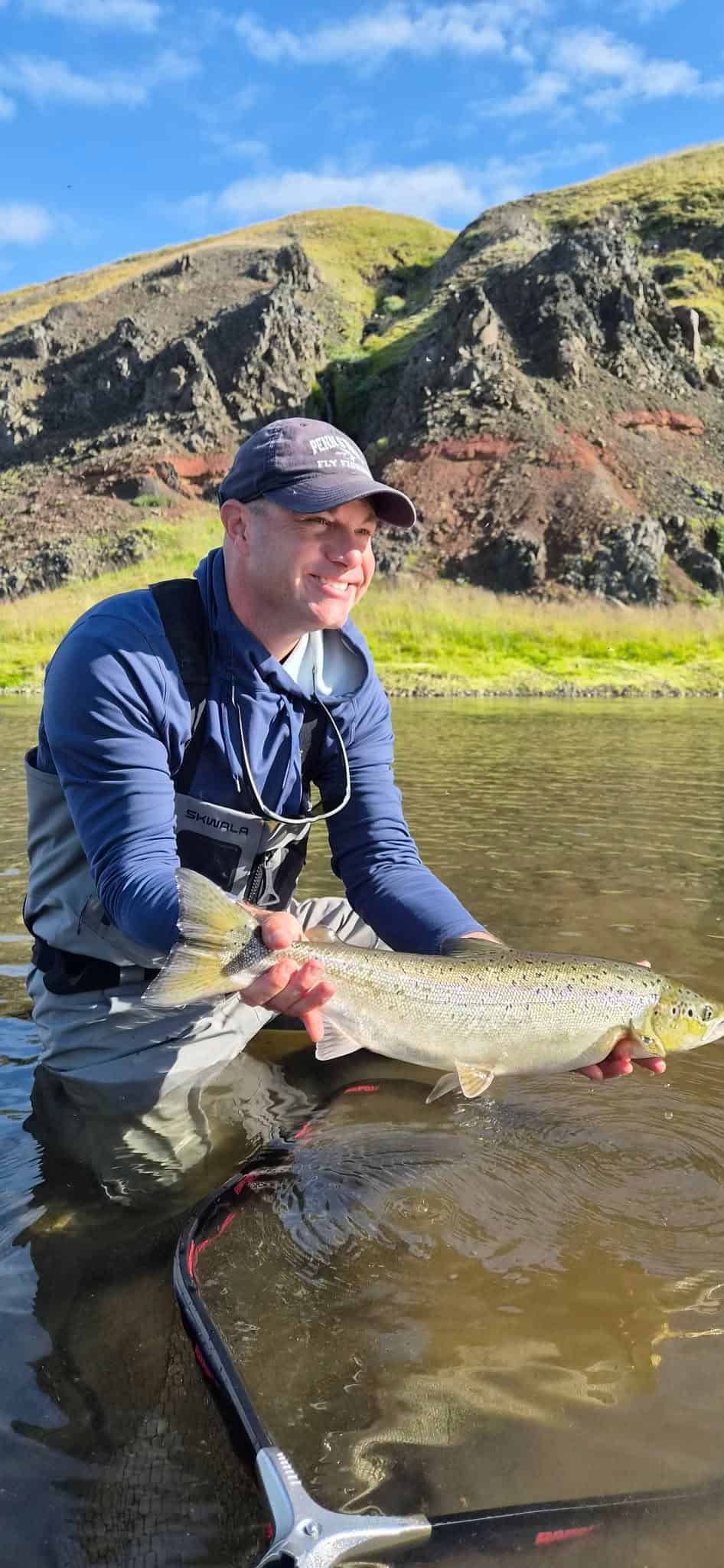Fisherman holding a large fish in a river with green hills in the background.