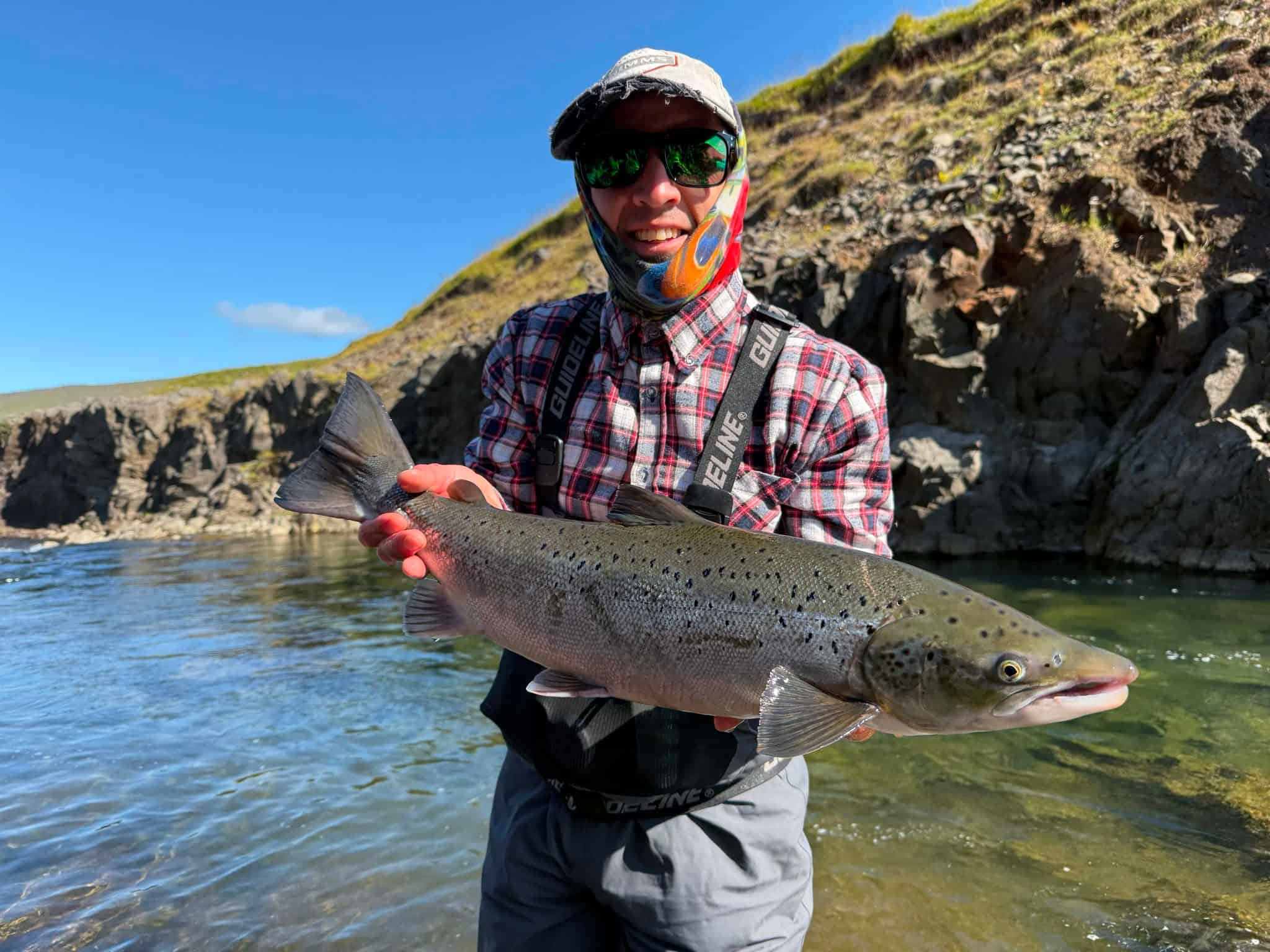 Fisherman holding a large fish in a river with cliffs in the background.