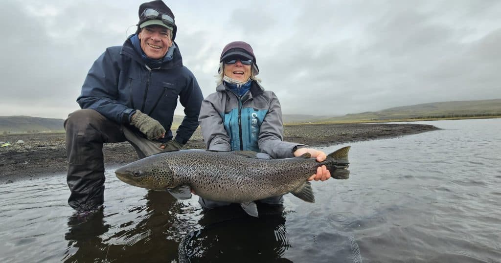 Real people fishing and holding a large rainbow trout on a riverbank under cloudy skies.