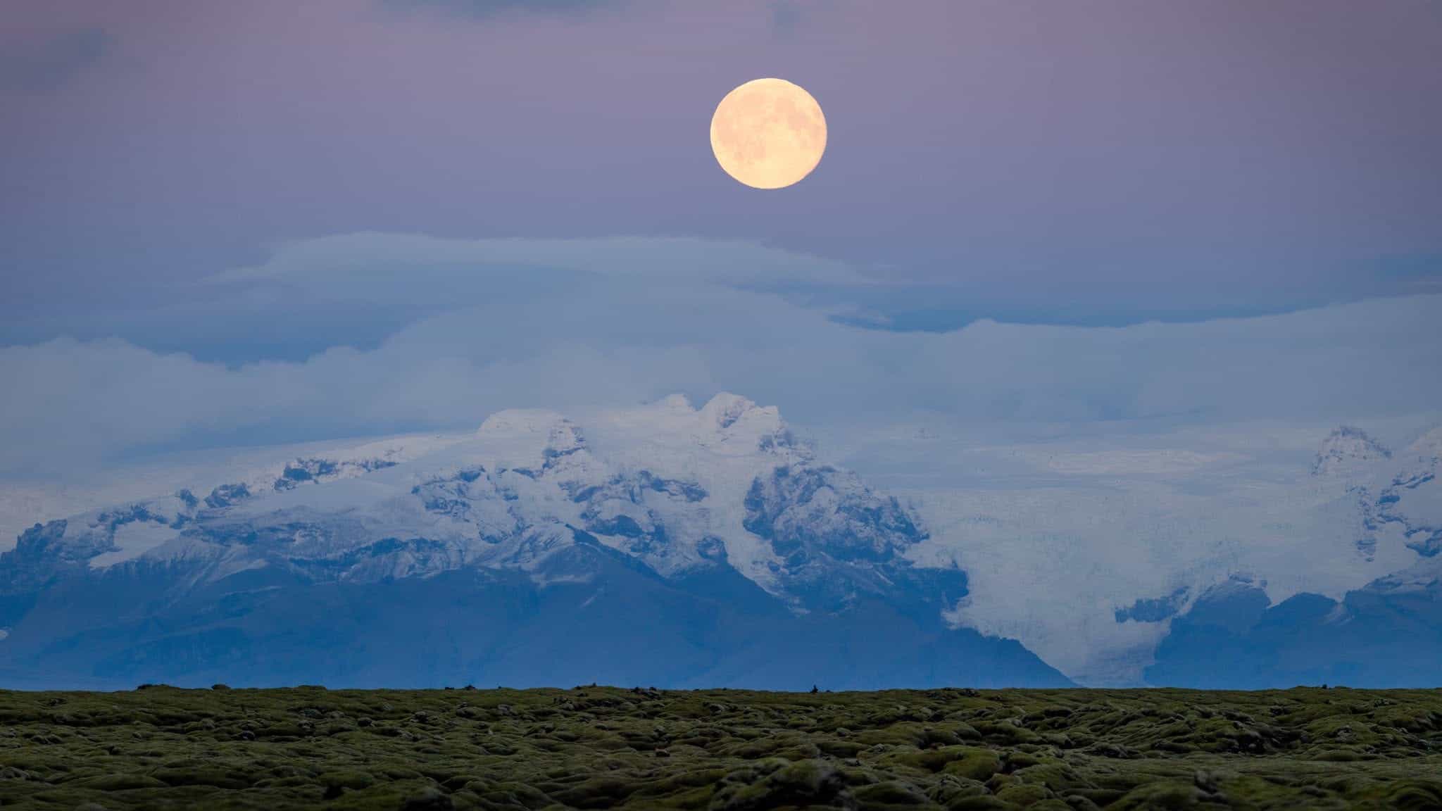 Full moon over snow-capped mountains and lush green landscape at dusk.