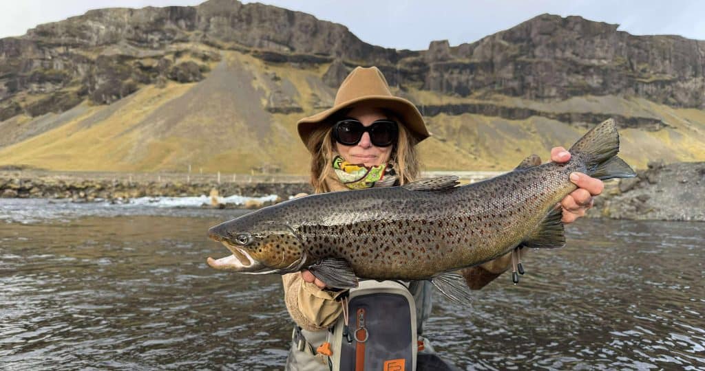 Large rainbow trout caught during fly fishing in river with scenic mountain background.