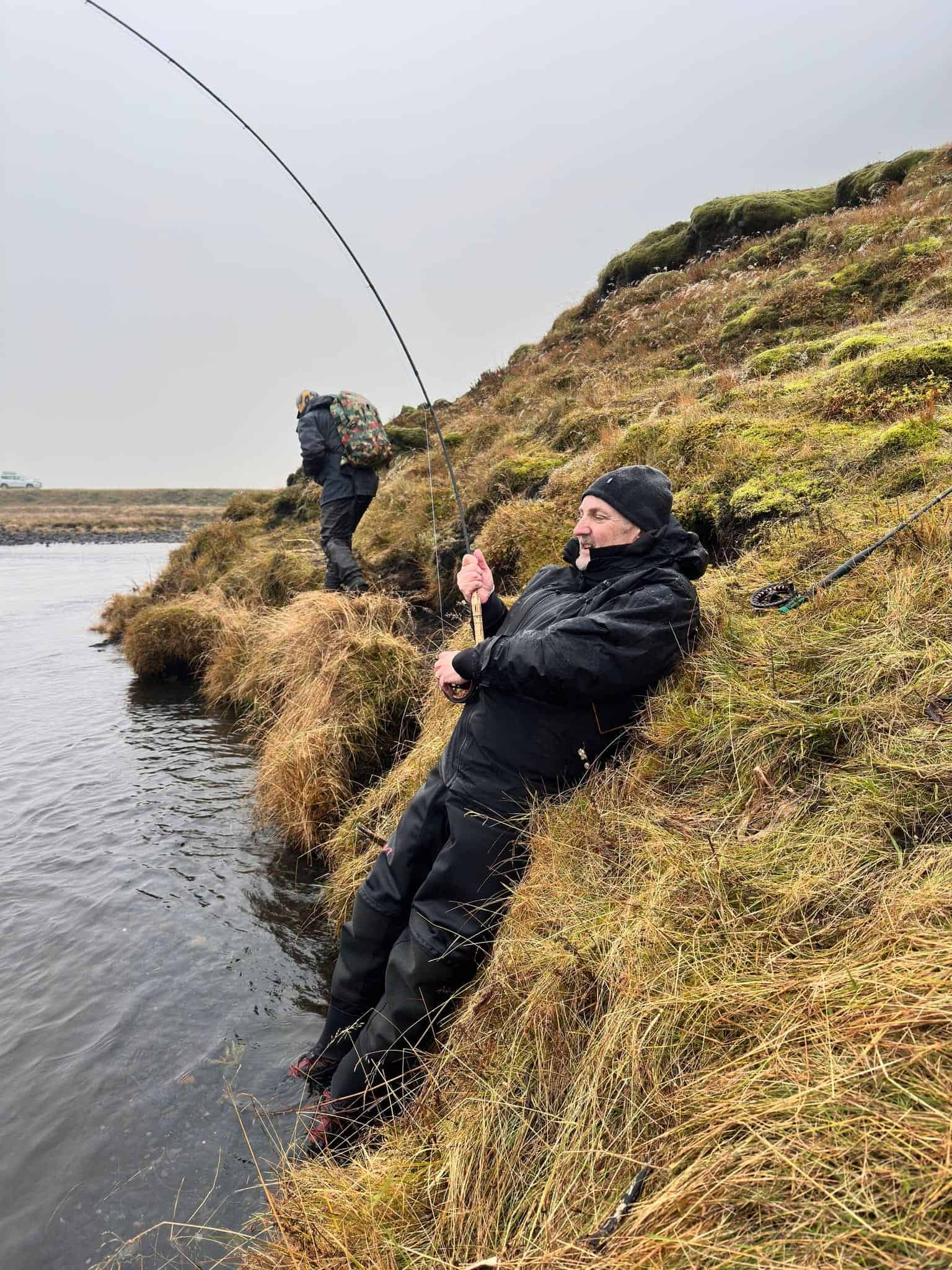 Fishing on riverbank with anglers in outdoor gear near water, overcast weather, fishing equipment, and natural landscape in background.