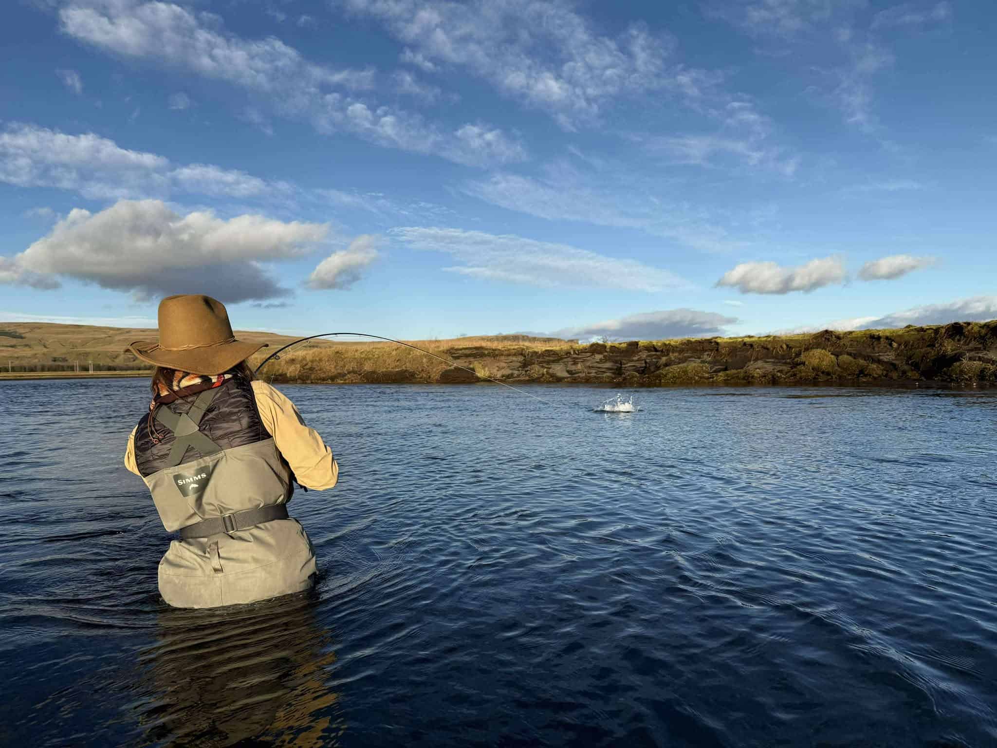 Fishing enthusiast casting line in river with scenic landscape and blue sky.