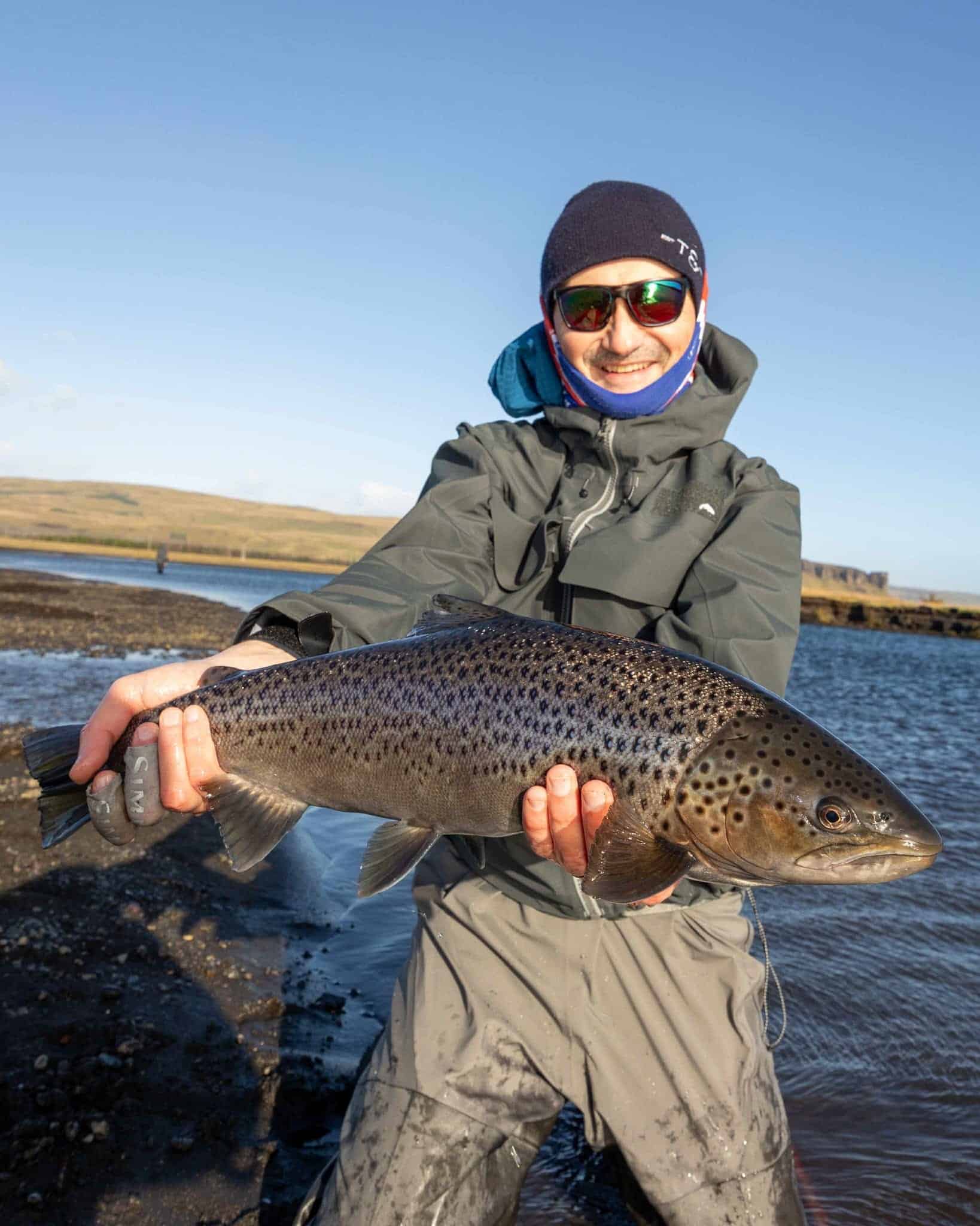 Vibrant image of a man holding a large, freshly caught rainbow trout by a river in Iceland. Perfect for fishing, outdoor adventures, and angler lifestyle content.