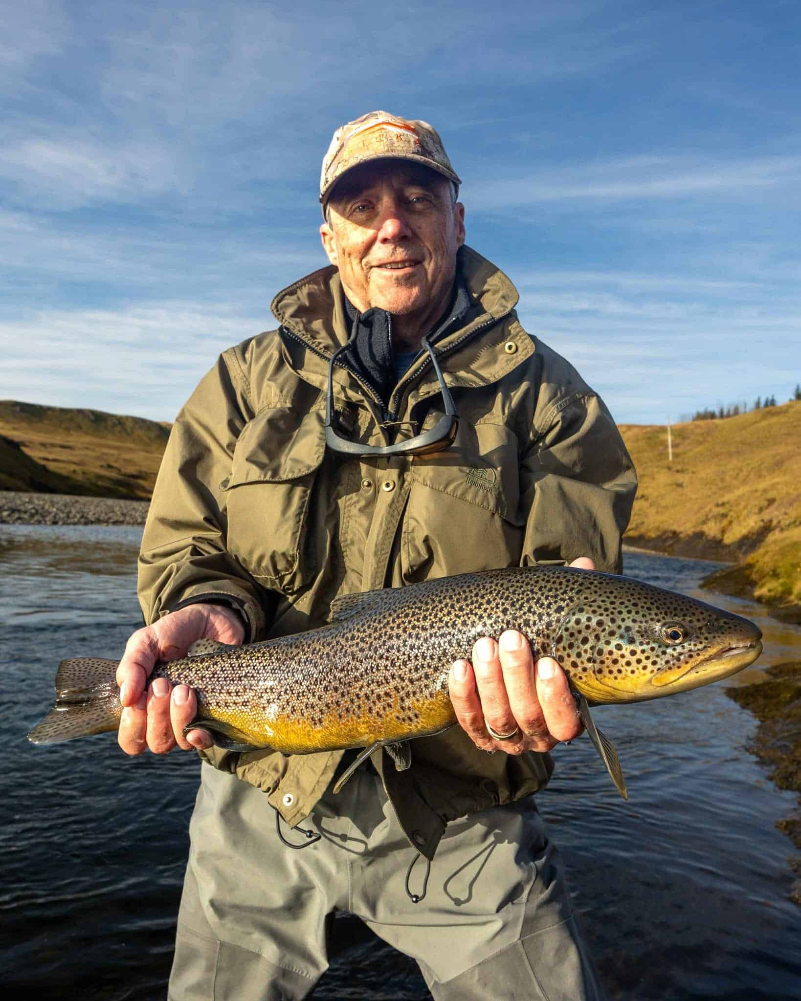 Vast catch of rainbow trout held by angler with a scenic river and hills in the background.