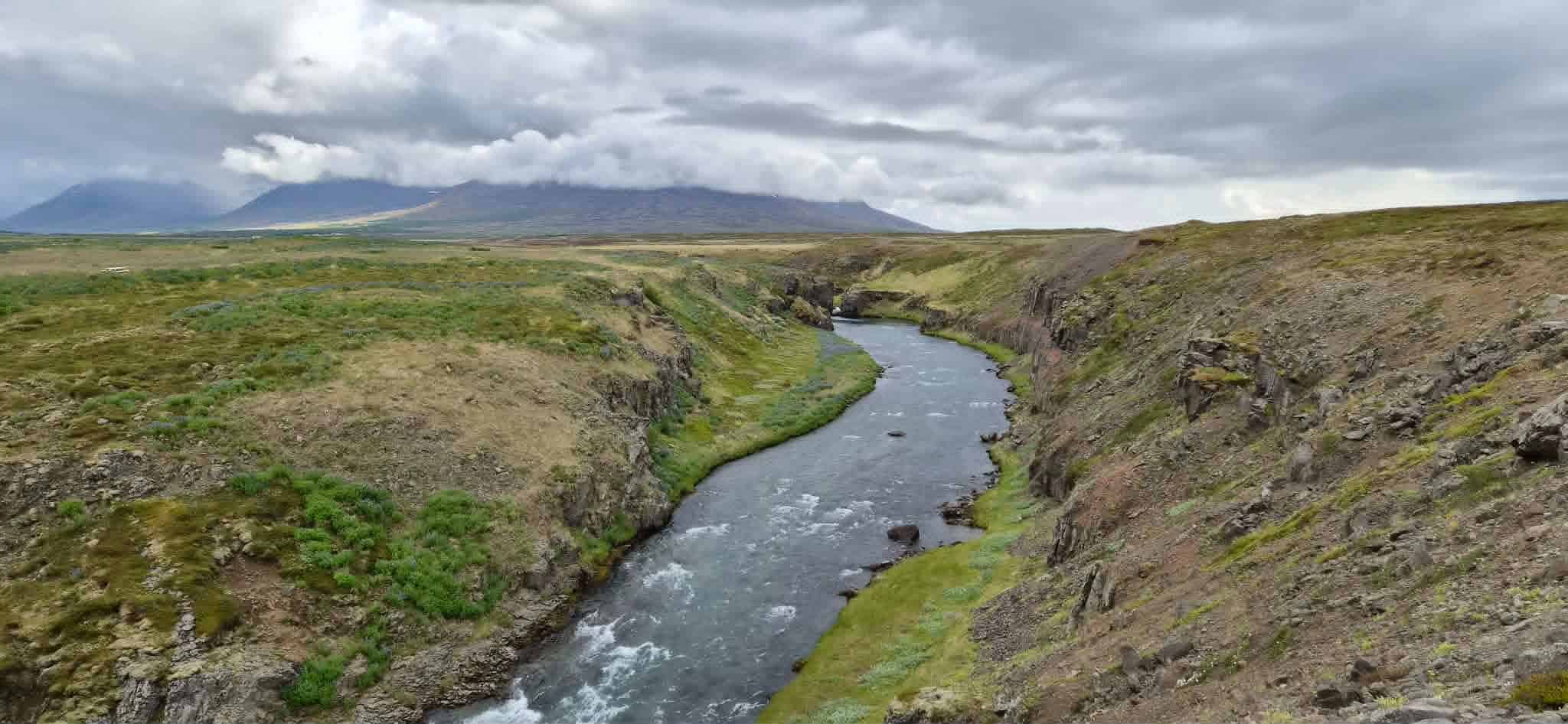 Wide view of a river flowing through a green valley with cloudy sky.