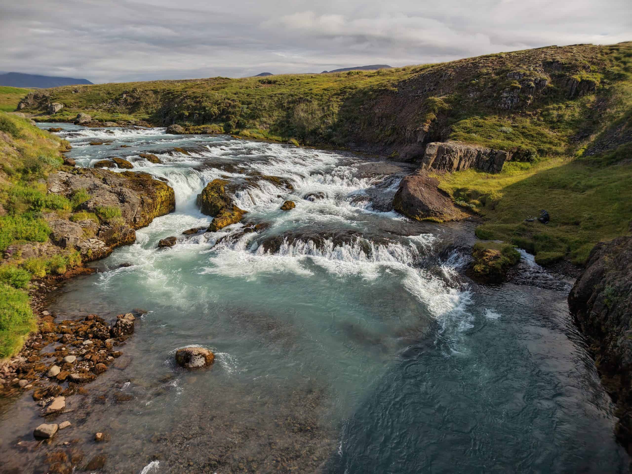 Scenic Icelandic river with rapids and green banks under cloudy sky.