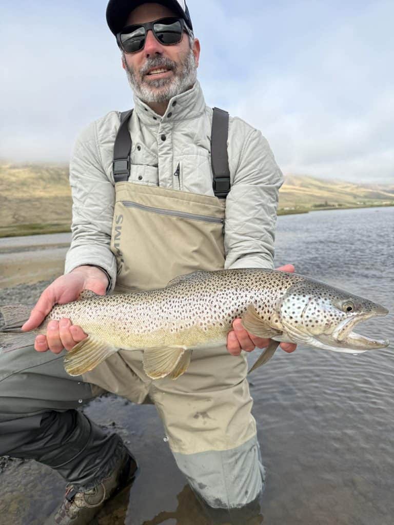 Angler holding a brown trout by a river with scenic background.