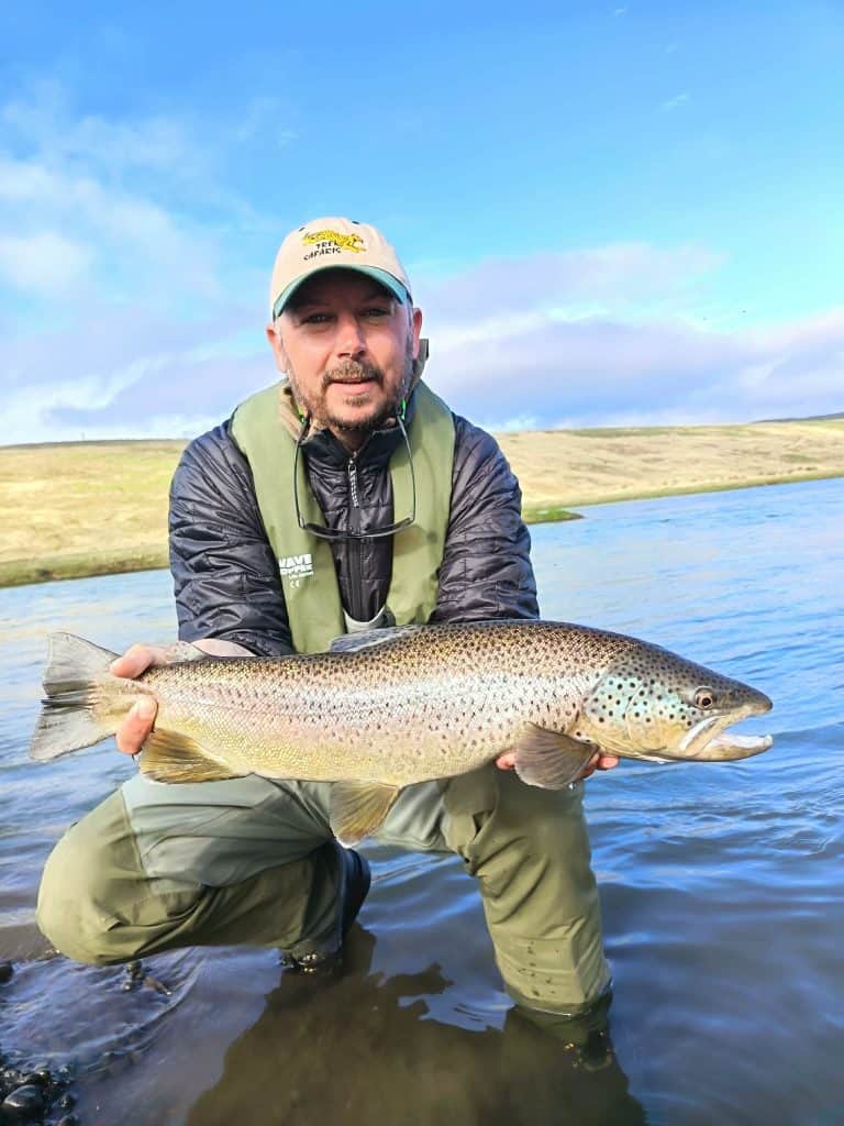 Angler holding a large brown trout in a river with blue sky.