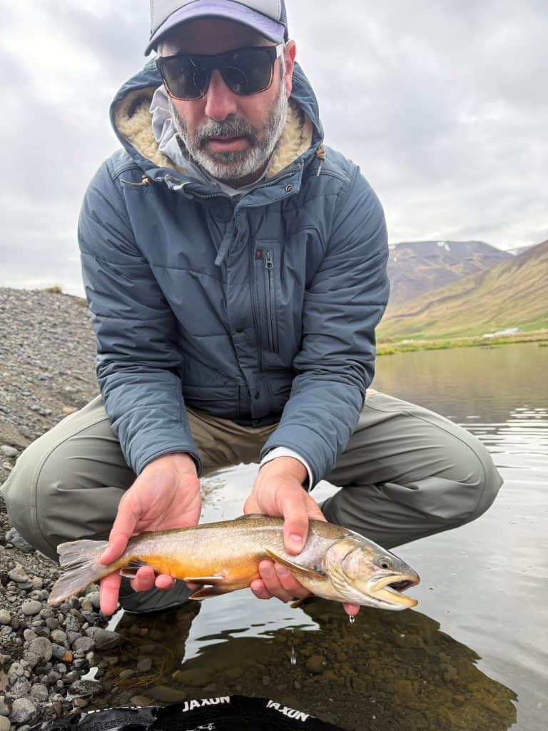 Man holding a fish by the water in Iceland, scenic mountain background.