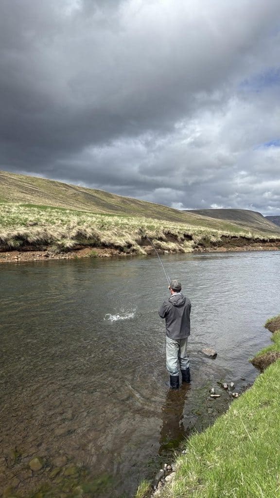 Fisherman casting line in a river with cloudy sky.