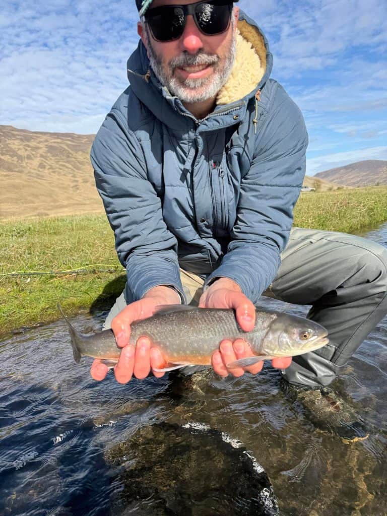 Man holding a fish in a river with mountains in background.