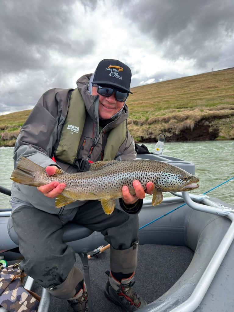Angler holding a brown trout in a boat on a river with cloudy sky.