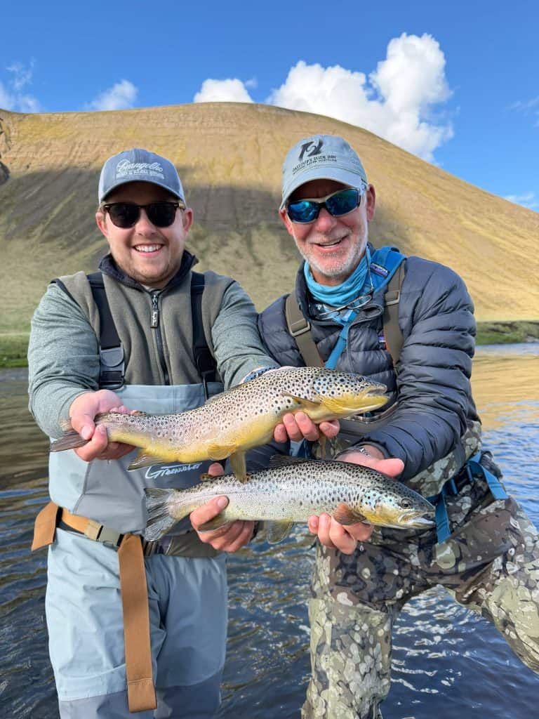 Two anglers holding a fish each in a scenic river setting.