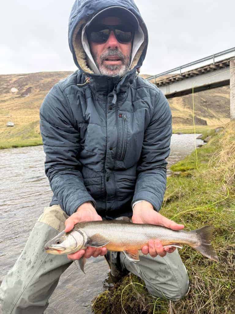 Man holding a sea run brown trout near a river with a bridge in the background.