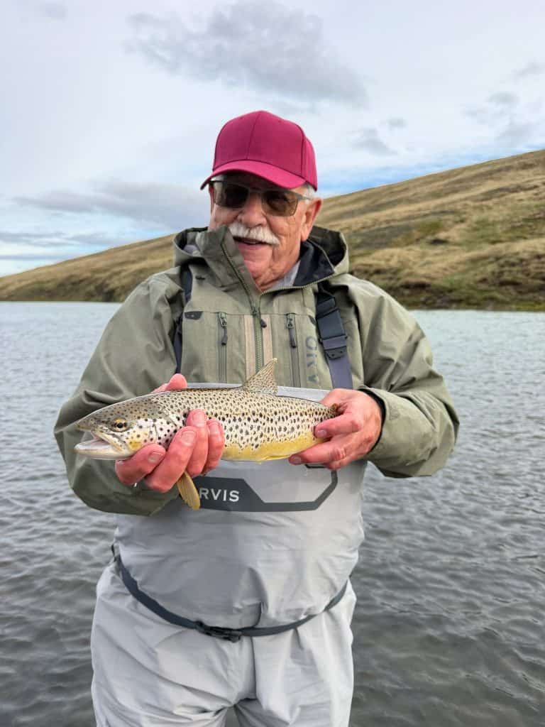 Elderly man holding a fish near a lake with hills in the background.