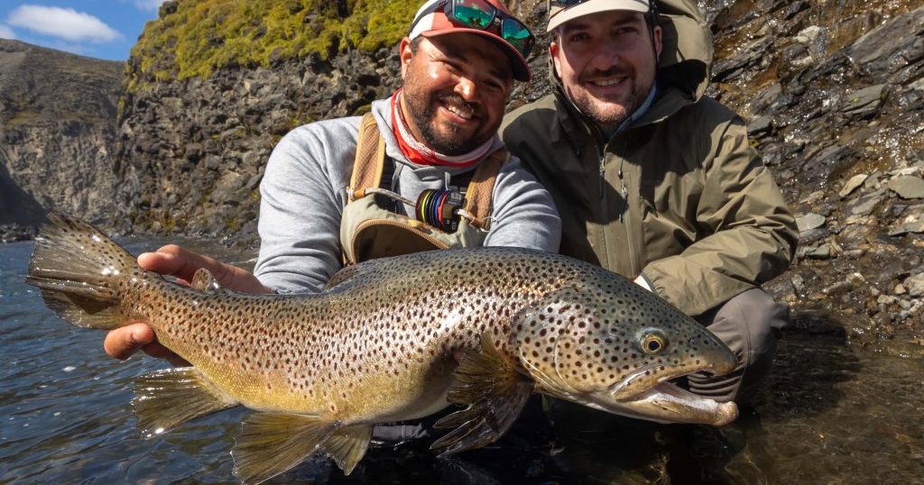 Two anglers holding a large brown trout in a river.