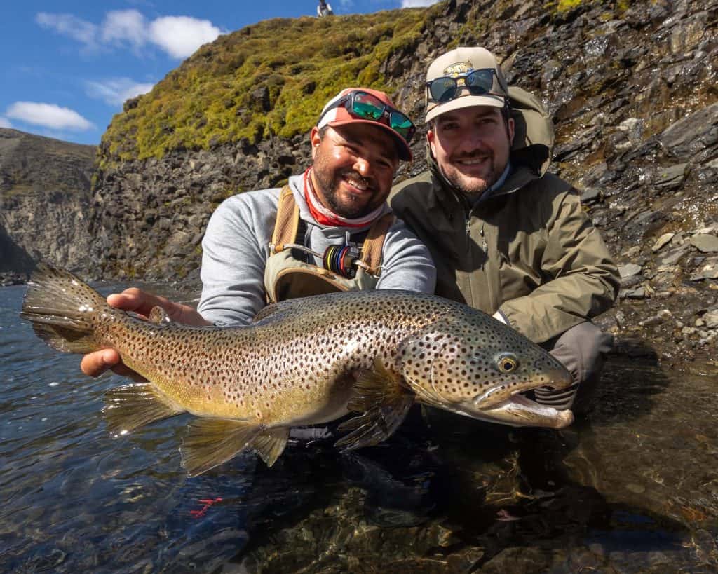 Two anglers holding a large brown trout in a river with scenic landscape.