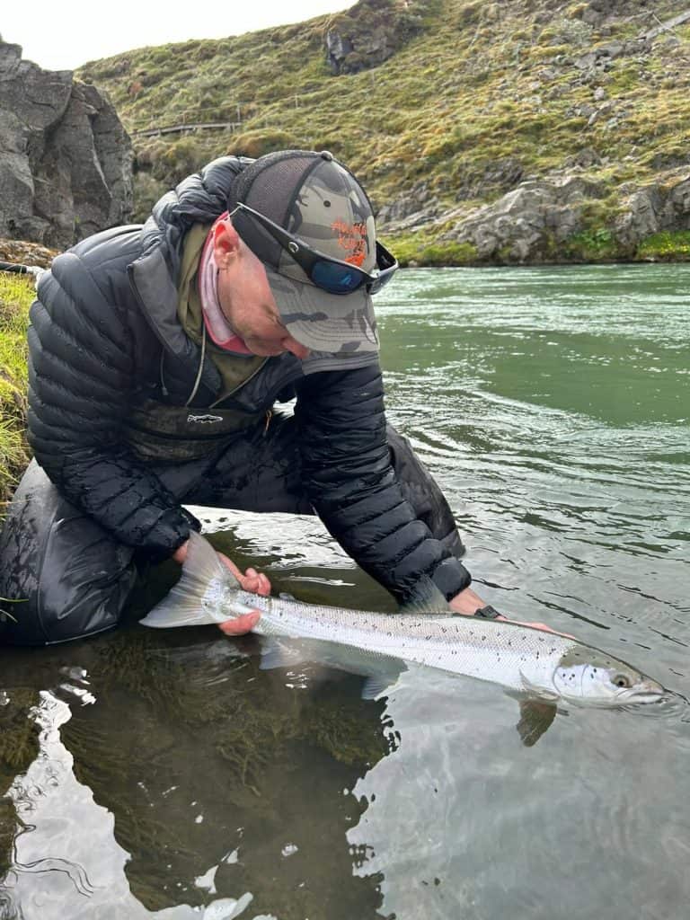 Angler holding a fish in a river with rocky banks and green hills.