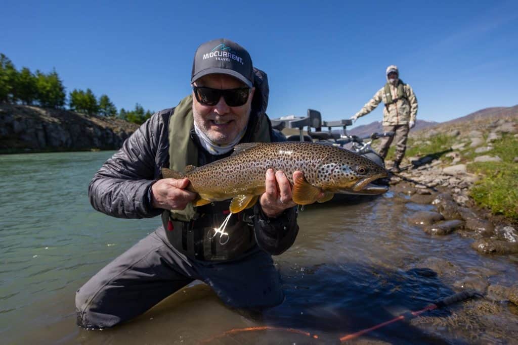 Angler holding a large brown trout in a river with scenic background.