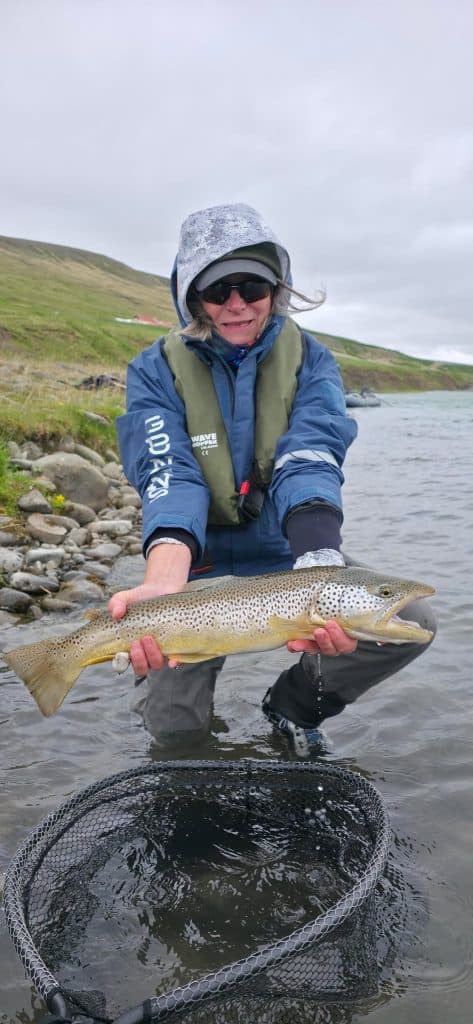 Angler holding a fish near a river in Iceland under cloudy sky.