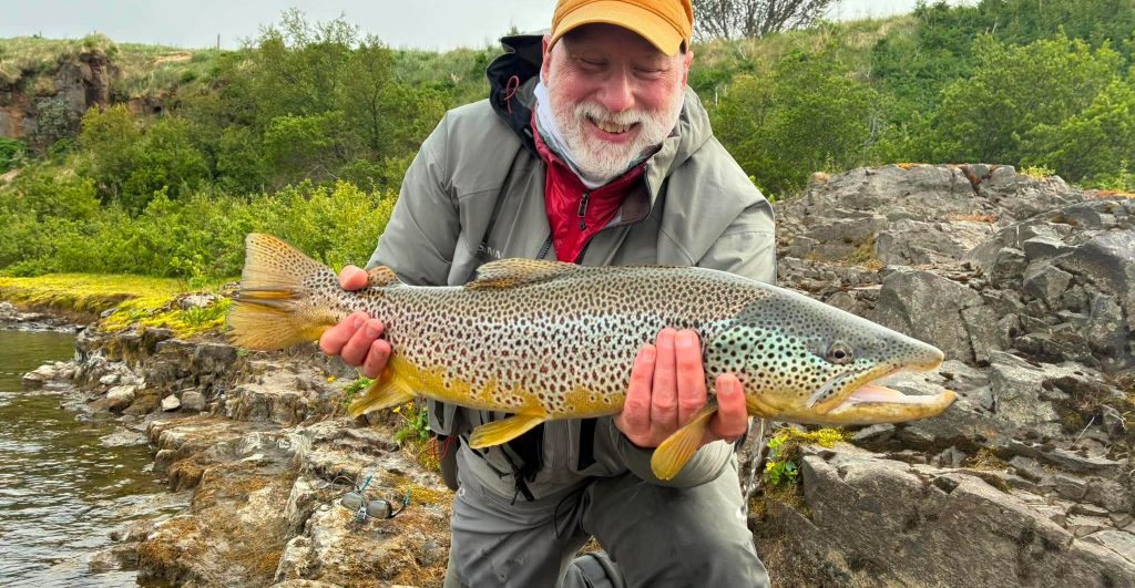 Angler holding a large brown trout near a river with rocks and greenery.