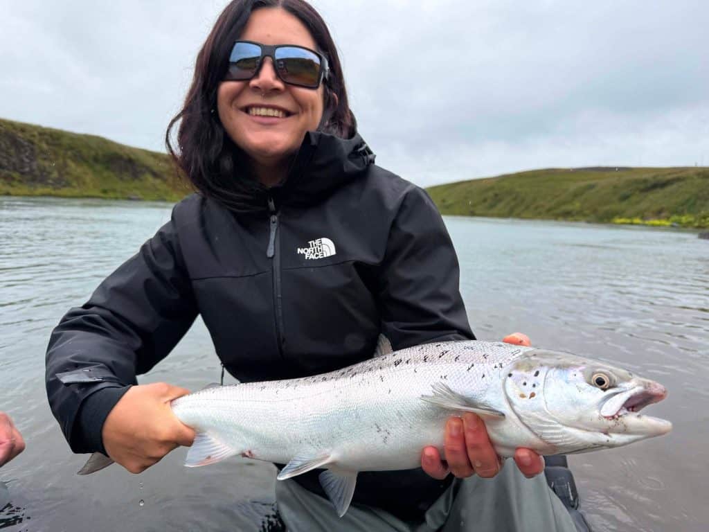 Woman holding a fish by a river with green hills in the background.