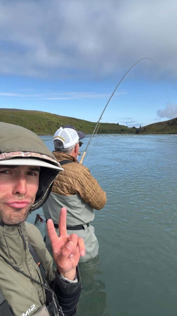 Two anglers fishing in a river with scenic hills in the background.