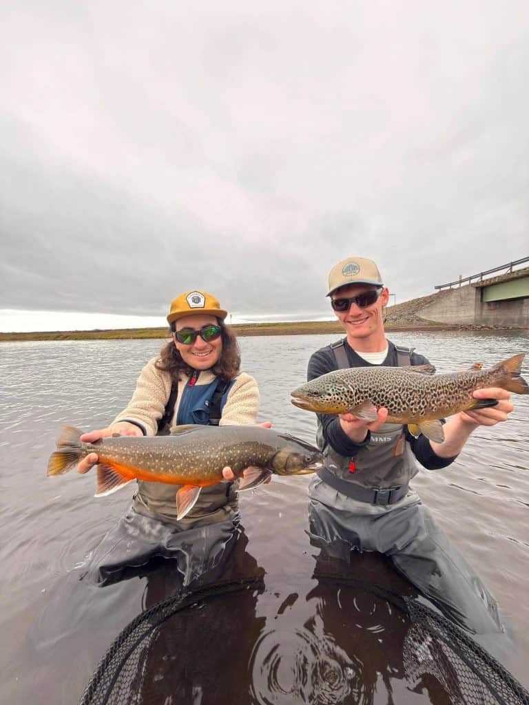 Two anglers holding fish in a river with a bridge in the background.