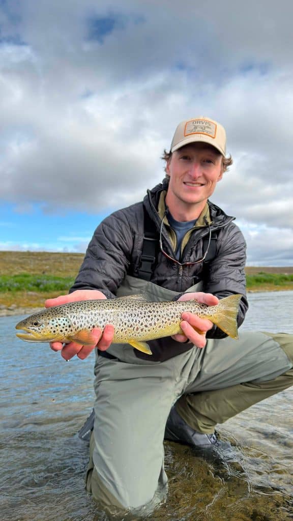 Angler holding a brown trout in Icelandic river with cloudy sky.
