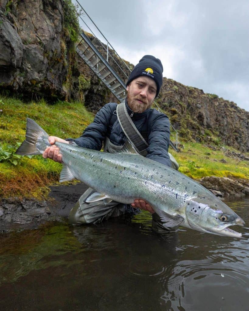 Man holding a large fish near a river with rocky cliffs in the background.