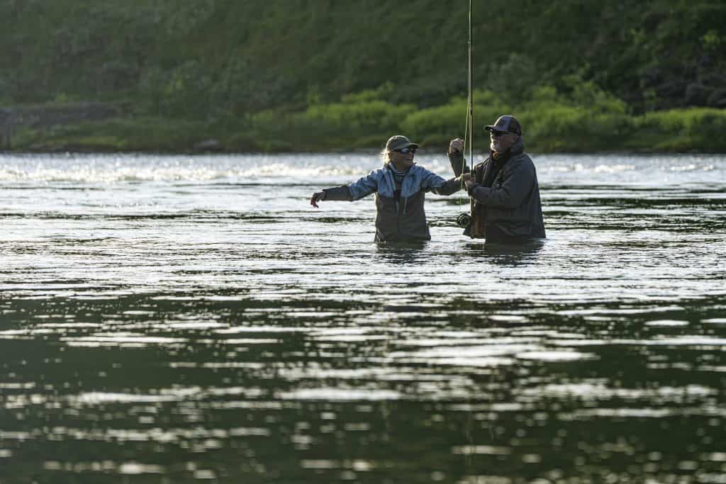 Two anglers fishing in a river during daytime with lush green surroundings.