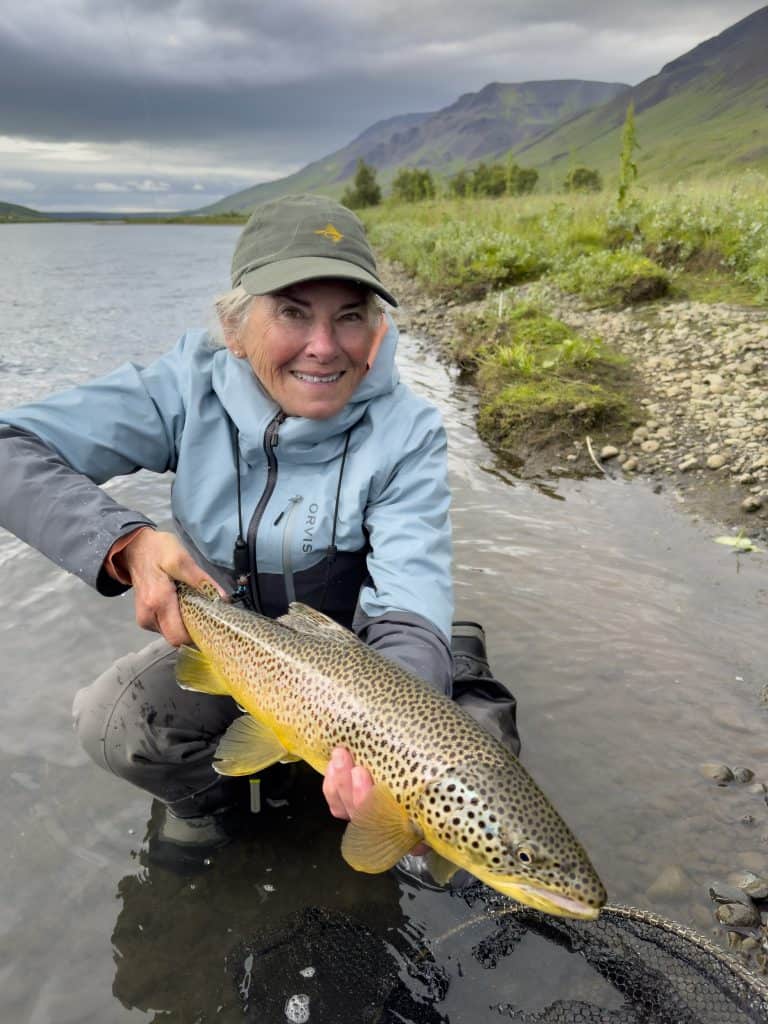 Smiling woman holding a brown trout by a river in Iceland.
