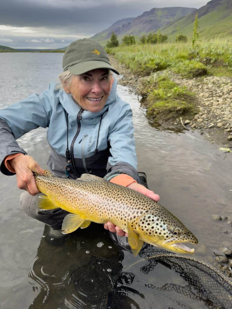 Smiling woman holding a brown trout by a river.