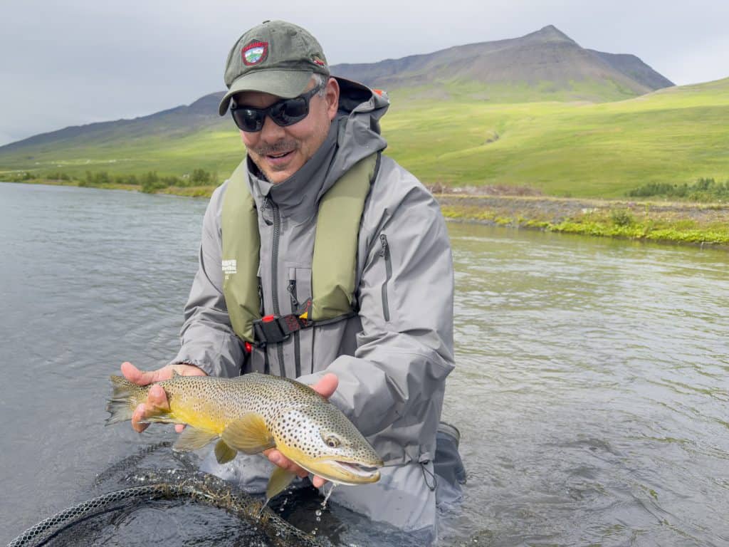 Man holding a fish in a river with mountains in the background.