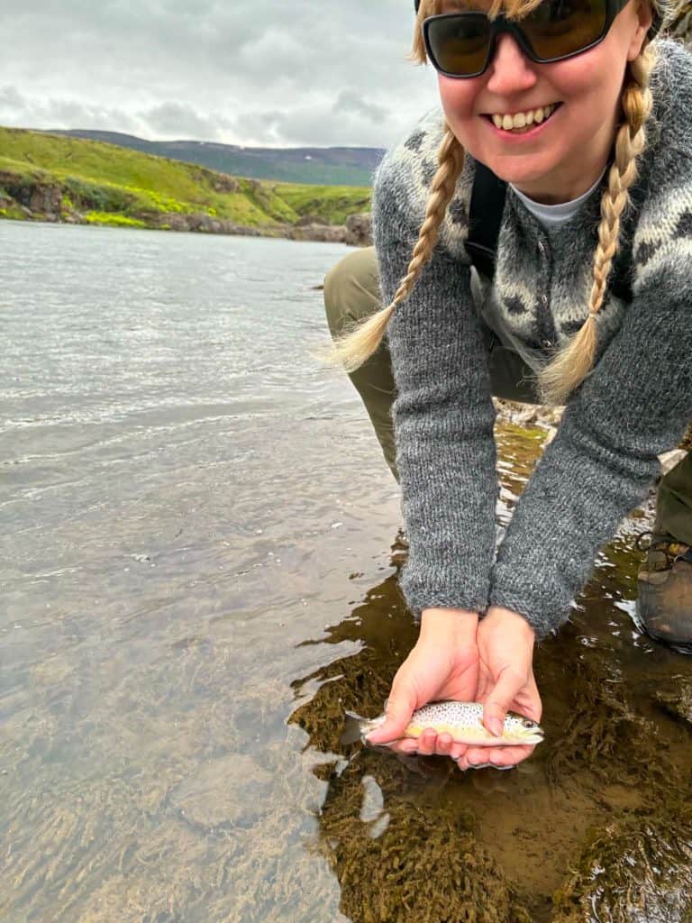 Woman holding a fish by the riverbank during fishing trip.