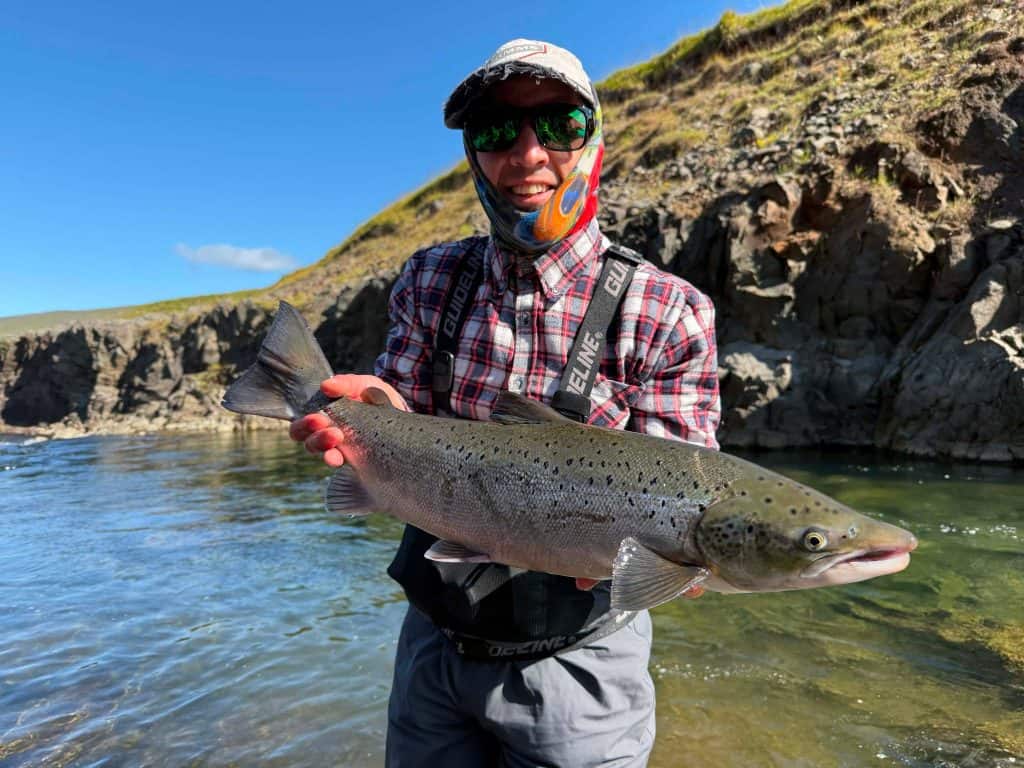 Angler holding a large fish near a river with cliffs in the background.