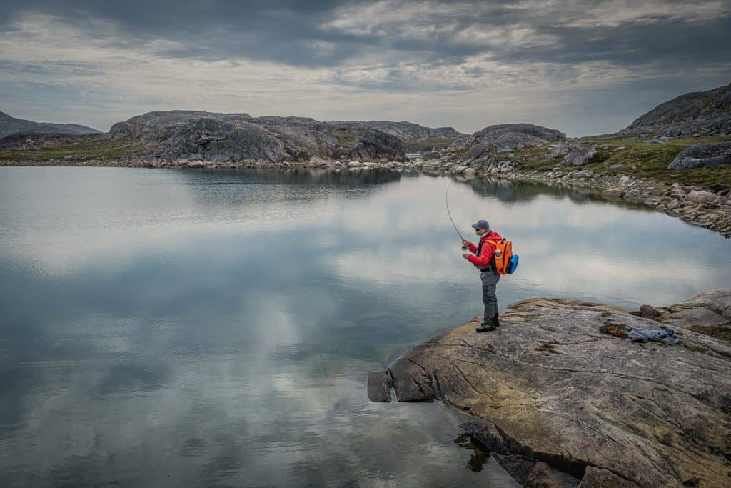 Fly fisherman casting a line from a rocky shoreline in a remote mountain lake.