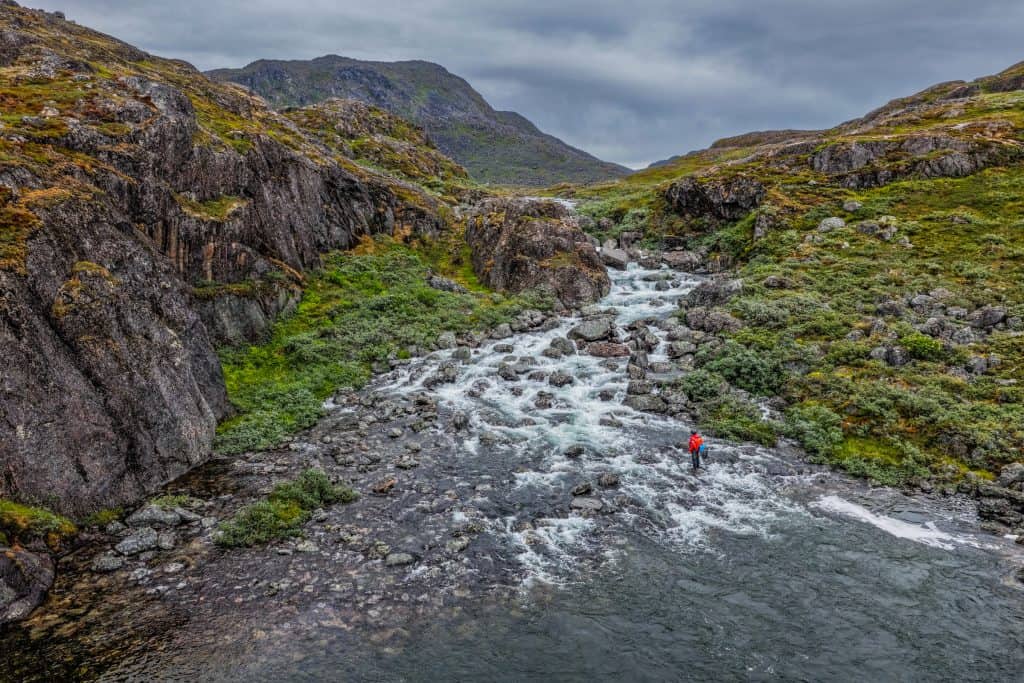 Fly fisher wading in a rugged Icelandic river surrounded by mossy rocks and mountains.