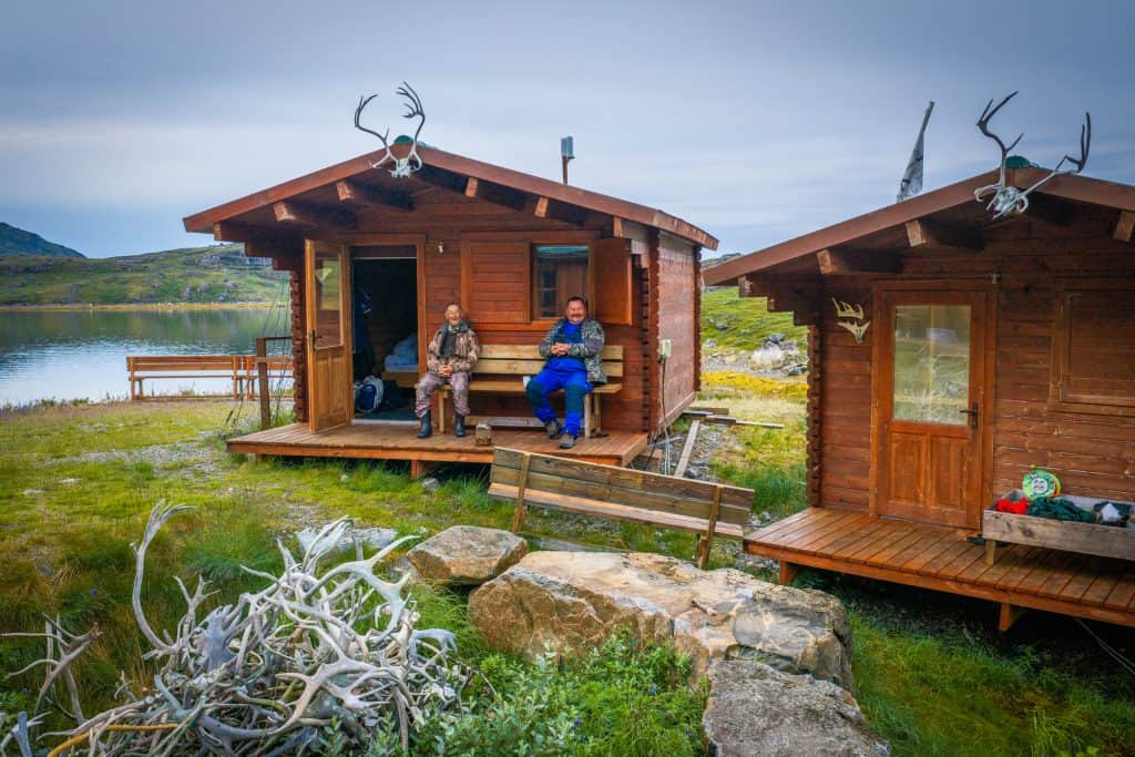 Two men sitting on the porch of a wooden lodge with antlers on the roof, overlooking a lake.