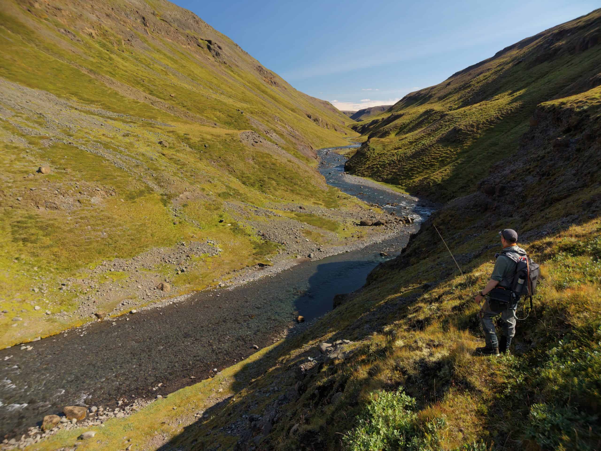Fly fisherman casting in Icelandic river surrounded by green hills.