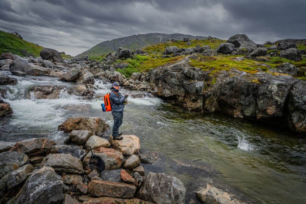 Fly fisherman casting in a rocky river with cloudy sky overhead.