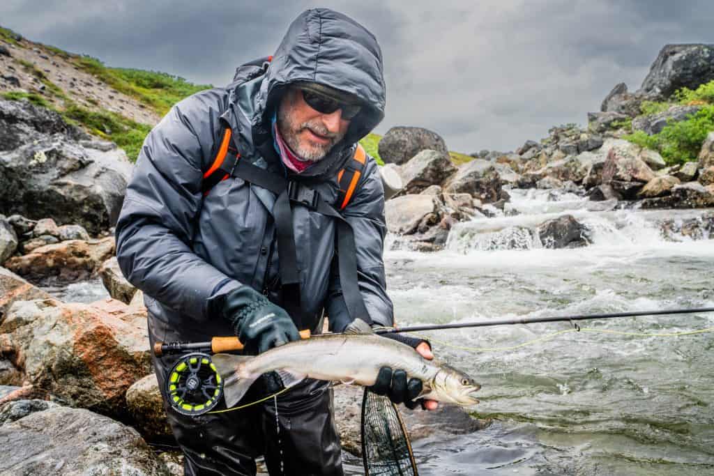 Fisherman holding a fish in a river with rocks and cloudy sky.